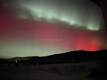 Esta imagen muestra la aurora boreal sobre un paisaje oscuro. Auroras verdes y rojas iluminan el cielo nocturno sobre colinas lejanas y luces dispersas.