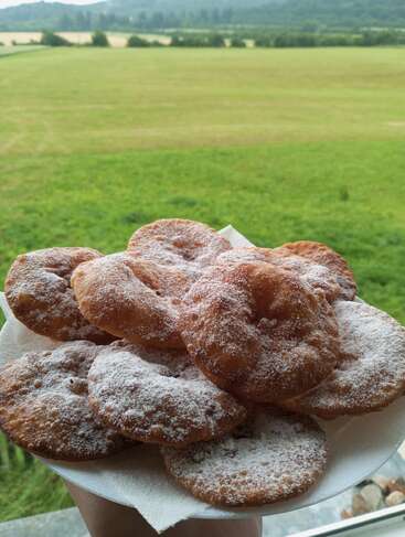 Um prato de bolinhos redondos recém-fritos, generosamente polvilhados com açúcar de confeiteiro, servido ao ar livre com um cenário pitoresco de campos verdes exuberantes e colinas ao longe.