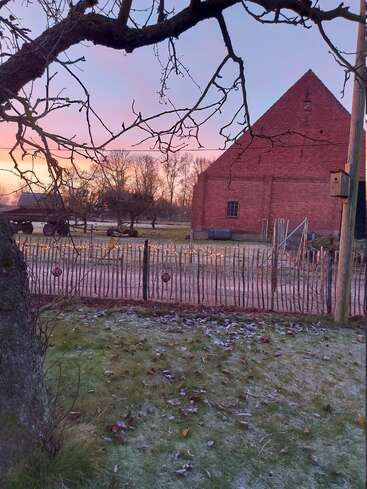 A frosty morning scene shows a rustic wooden fence, a red brick barn, bare tree branches, and soft pastel skies at sunrise in a quiet countryside.