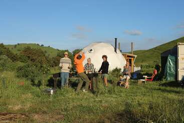 Five people and a child gather outdoors near a geodesic dome, enjoying sunny weather, greenery, and a picnic. The scene feels relaxed and joyful.