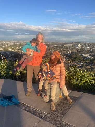 A family of four enjoys a sunny day on a hilltop, overlooking a city. The parents and children wear colorful jackets, smiling warmly together.