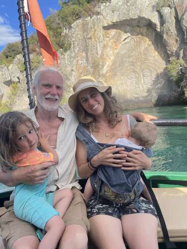 A smiling family of four poses on a boat, with scenic rock carvings in the background. The mother holds a baby, the father hugs their daughter.
