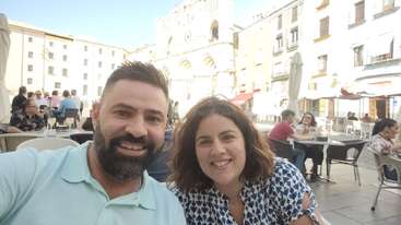 A smiling man and woman sit together at an outdoor café in a lively plaza, with historic buildings and people enjoying the sunny, relaxed atmosphere in the background.
