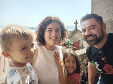 A smiling family of four poses together outdoors. In the background, there’s a large cross, rooftop, and a scenic view under a clear, sunny sky.