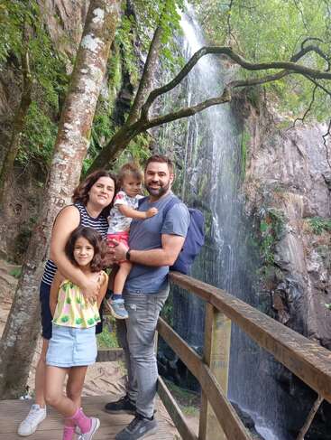 A happy family of four poses on a wooden bridge in front of a beautiful waterfall, surrounded by lush green trees and rocky natural scenery.