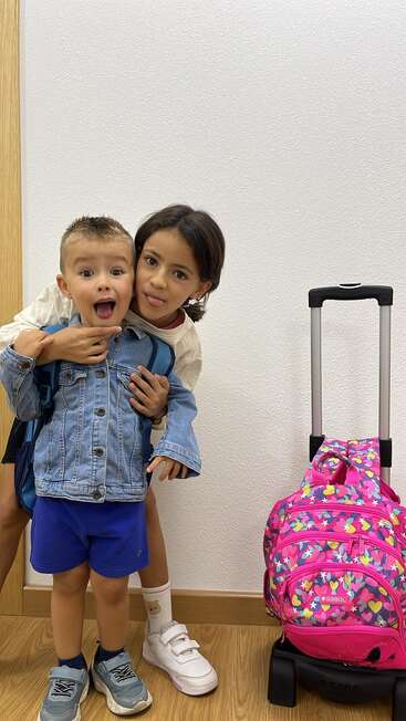 Zwei Kinder, ein Junge in Jeansjacke und ein Mädchen in Weiß, die beide die Zunge herausstrecken, stehen verspielt neben einem bunten rosa Rollrucksack.