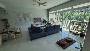 Das helle Wohnzimmer hat blaue Sofas, eine Weltkarte, große Fenster, Zimmerpflanzen, einen Esstisch, einen Balkon mit Pflanzen, einen Deckenventilator und Meditationskissen.