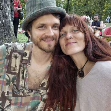 Una pareja sonriente posa muy junta al aire libre, rodeada de árboles y gente. El hombre lleva una camisa estampada y un sombrero; la mujer tiene el pelo largo y rojo y lleva un collar.