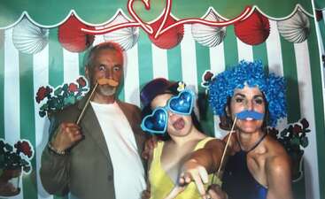 Trois personnes s'amusent dans un photomaton, vêtues de costumes ridicules : fausses moustaches, grosse perruque bleue, lunettes en forme de cœur et expressions enjouées sur fond de fête.