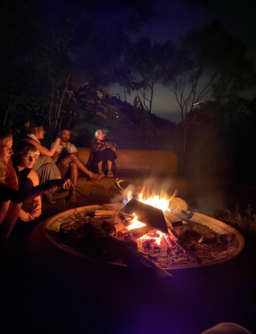 La imagen muestra a un grupo de personas reunidas en torno a una hoguera, con una gran olla suspendida de un trípode sobre las llamas, sobre un fondo oscuro.