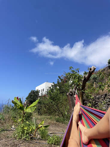 Eine Person entspannt sich in einer bunten Hängematte und genießt den klaren blauen Himmel, üppige grüne Pflanzen, ein weißes Gebäude und die friedliche Natur im Hintergrund.