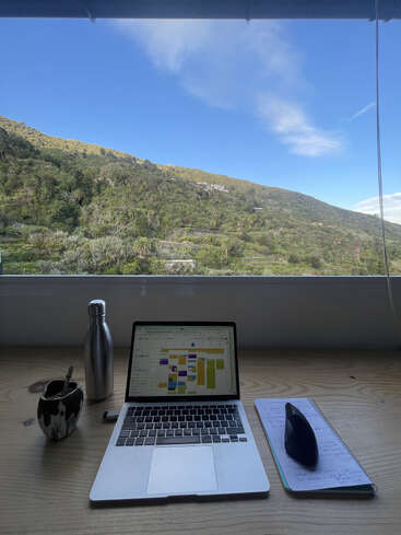 Ein Laptop steht auf einem Holztisch mit einem Notebook, einer Maus, einer Wasserflasche und einem Becher. Ein helles Fenster gibt den Blick auf die grünen Hügel und den blauen Himmel frei.