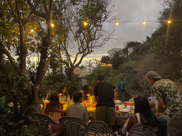Eine Gruppe von Menschen versammelt sich in der Abenddämmerung im Freien und teilt ihr Essen an einem Tisch unter Lichterketten, umgeben von Bäumen, die eine warme und festliche Atmosphäre schaffen.