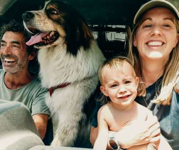 Une famille heureuse est assise dans une voiture, le sourire aux lèvres. Un gros chien au pelage soyeux est assis entre un homme et une femme, tandis que celle-ci tient un jeune enfant dans ses bras.