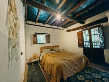Cette chambre confortable est dotée d'un plafond rustique en bois, d'un lit double avec une literie couleur beige, d'une petite table de chevet, d'une lumière naturelle provenant des fenêtres, de murs blancs et d'une décoration murale de style bohème.