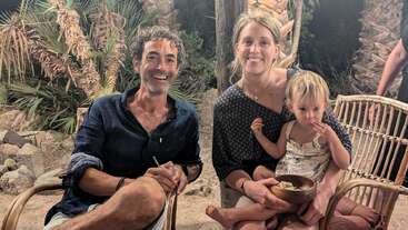 Un homme, une femme et un jeune enfant sont assis en plein air sur des chaises en osier, tout sourire, entourés de palmiers. L'enfant tient un bol, et l'ambiance semble chaleureuse et détendue.
