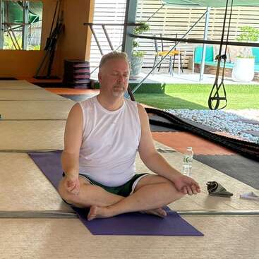 Un homme est assis en tailleur sur un tapis de yoga à l'intérieur, vêtu d'une chemise blanche et d'un short vert, les yeux fermés, méditant paisiblement avec une bouteille d'eau à proximité.