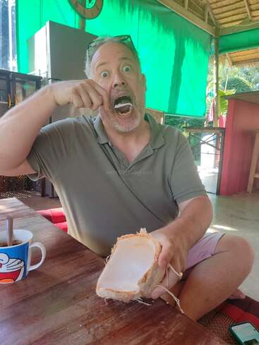 Un hombre con expresión de sorpresa come con entusiasmo coco fresco con una cuchara, sosteniendo la cáscara del coco, sentado en una mesa de madera en una cafetería informal al aire libre.