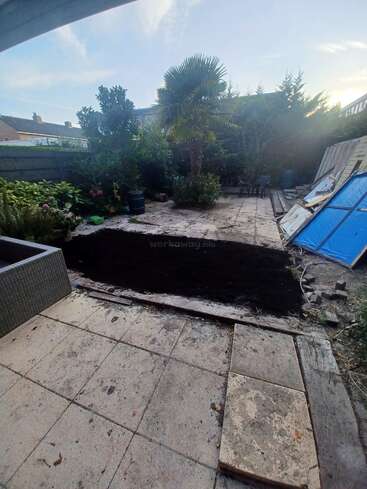 Esta imagen muestra un patio trasero con una parte de las baldosas del patio retirada, dejando al descubierto la tierra. Hay árboles, plantas, muebles de exterior y materiales de construcción cerca. El cielo está despejado.