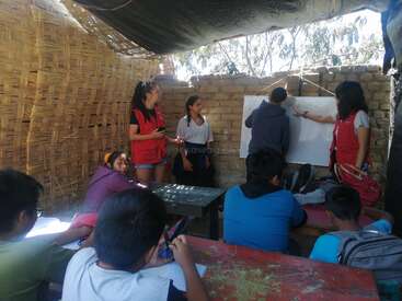 In a rustic outdoor classroom with woven walls, children attentively watch as teenagers teach and write on a whiteboard, creating a lively and engaging learning environment.