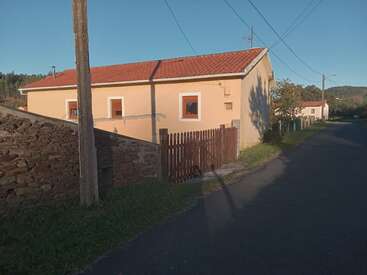 Una pintoresca casa con tejado rojo se alza junto a una carretera tranquila. Un muro de piedra, una verja de madera, árboles y cables telefónicos completan este apacible paisaje rural.
