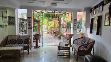 The image shows a cozy indoor lounge with wicker furniture, glass doors opening to a sunny garden patio, shaded seating, plants, and a relaxed, welcoming ambiance.