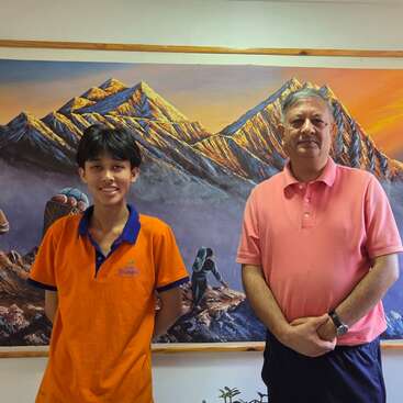 A young boy and an older man stand in front of a colorful mountain painting, both smiling, wearing orange and pink shirts, relaxed and friendly atmosphere.