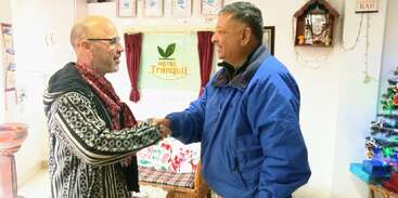 Two men are shaking hands and smiling inside a hotel lobby. The background has a “Hotel Tranquil” sign and festive decorations, creating a warm, welcoming atmosphere.