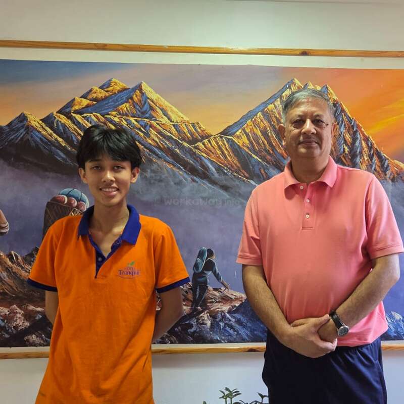 A young boy and an older man stand in front of a colorful mountain painting, both smiling, wearing orange and pink shirts, relaxed and friendly atmosphere.