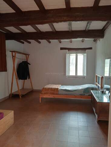 Dieses Bild zeigt ein schlichtes, rustikales Schlafzimmer mit sichtbaren Holzbalken, einem Einzelbett, einem Kleiderständer aus Holz, einer Kommode mit Spiegel und einem Fliesenboden.