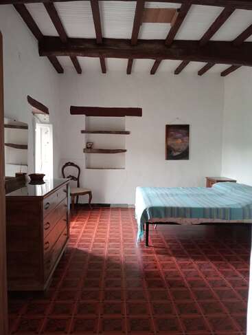 Das Bild zeigt ein rustikales Schlafzimmer mit Holzbalken, Fliesenboden, einem schlichten Bett, einer Holzkommode, einem Stuhl, offenen Regalen und einem Gemälde an einer weißen Wand.