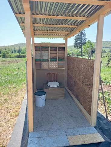 Dieses Bild zeigt eine einfache Komposttoilette im Freien mit einem Holzrahmen, einem Metalldach und offenen Seiten, eingebettet in eine ländliche, grasbewachsene Landschaft mit Bäumen.