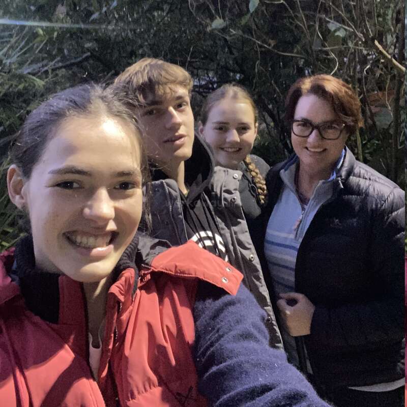 Four people stand close together outdoors, smiling for a selfie. They are wearing warm jackets, surrounded by greenery in the background, enjoying a casual, happy moment.