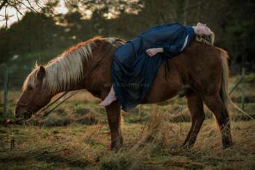 A woman in a long blue dress lies back on a brown horse, in a peaceful field, with trees and soft sunset light surrounding them.