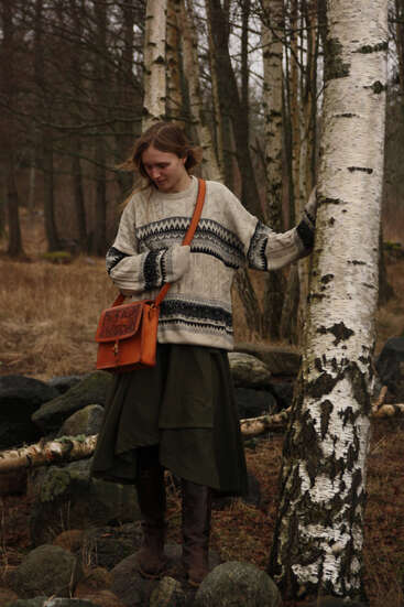 A woman stands in a forest among birch trees, wearing a patterned sweater, long skirt, boots, and carrying a brown leather bag, looking down thoughtfully.