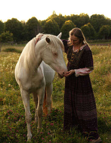 A woman in a long, patterned dress gently pets a white horse in a sunlit meadow, surrounded by wildflowers and trees on a peaceful evening.