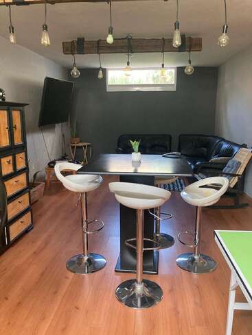 The room features wood flooring, modern bar stools, a square table, hanging light bulbs, black couches, a flat-screen TV, plants, and a casual, cozy atmosphere.