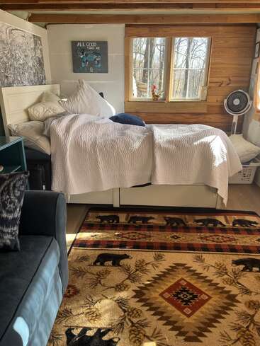 A cozy rustic bedroom with a white bed, lots of pillows, a bear-patterned rug, sunlight streaming in, wood walls, a window, and simple decor.