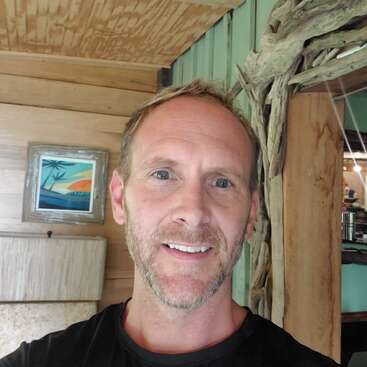 A man with short, light brown hair and a trimmed beard smiles indoors. Behind him, wooden walls, a framed tropical painting, and rustic decor create a cozy atmosphere.