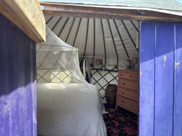 Através de uma porta azul, revela-se o interior de uma yurt aconchegante, com uma cama com dossel branco, cômoda, roupas penduradas, janela circular e carpete.
