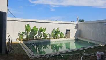 Esta imagen muestra una pequeña piscina rectangular rodeada de muros de hormigón. Unas plantas verdes bordean uno de los lados, y una manguera y un cubo descansan cerca bajo un cielo azul despejado.