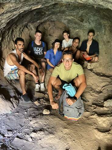 Das Bild zeigt sieben Personen, die in einer von Felsen umgebenen Höhle sitzen, mit einer Person, die davor hockt, und die alle in einer entspannten und lockeren Atmosphäre zu sein scheinen.