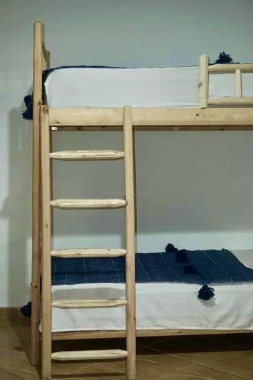 Das Bild zeigt ein helles Etagenbett aus Holz mit einer Leiter, weißen Laken und einer dunkelblauen Decke auf dem unteren Bett vor einer schlichten Wand und einem Holzboden.
