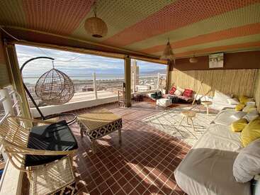 Das Bild zeigt ein geräumiges Zimmer mit Balkon, das mit einem Bett, einer Sitzecke und einem Hängesessel ausgestattet ist und einen malerischen Blick auf das Meer und die fernen Berge bietet.