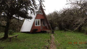 The image depicts a small, red A-frame house with a white roof, situated in a grassy yard, surrounded by trees and a cloudy sky.