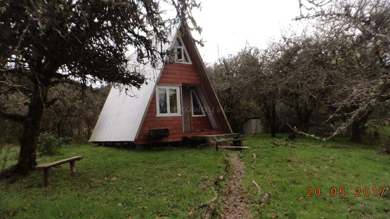 The image depicts a small, red A-frame house with a white roof, situated in a grassy yard, surrounded by trees and a cloudy sky.