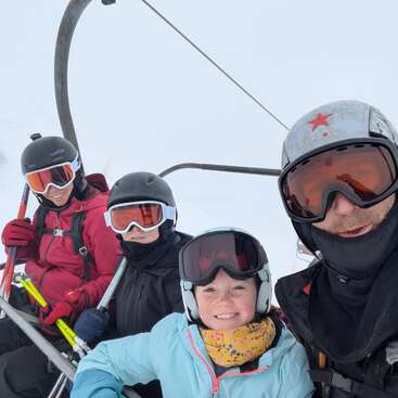 Four people wearing ski gear and helmets smile while sitting on a ski lift, surrounded by snow and cloudy skies, ready for a fun day of skiing.