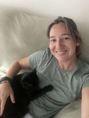 Uma mulher está sorrindo sentada no sofá, vestindo uma blusa verde. Um gato preto está deitado confortavelmente no peito dela, descansando tranquilamente enquanto ela o segura.