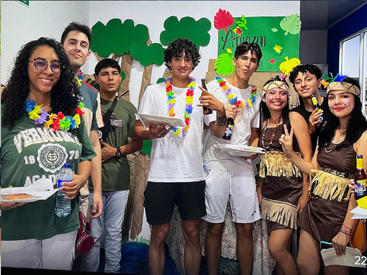 A group of young people pose together, wearing festive, Amazon rainforest-themed costumes and leis, holding food and drinks, smiling and celebrating in a decorated room.