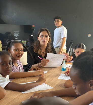 A group of children and a teacher sit around a table, engaged in an activity with worksheets and pencils. Another person stands in the background, observing.